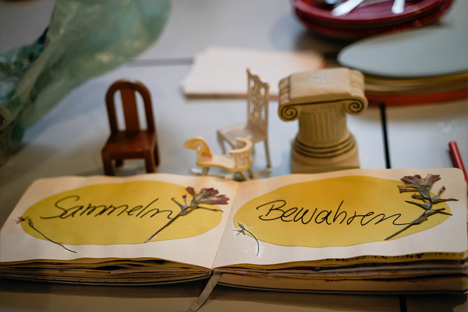 Aufgeschlagenes Notizbuch. Auf der linken Seite steht „Sammeln“ und auf der rechten „Bewahren“. Im Hintergrund stehen Miniatur-Möbel.