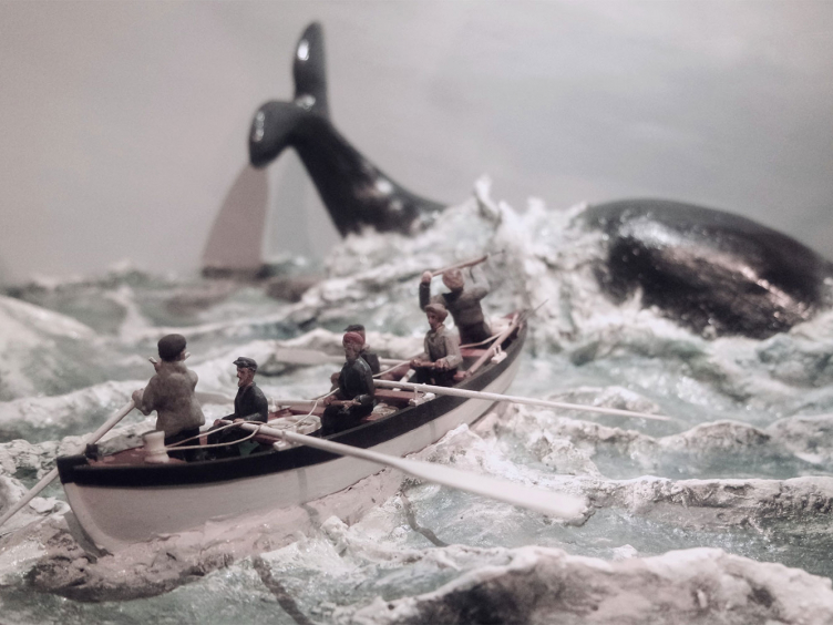 Ein Modell vom Walfang. Stürmisches Meer. Im Vordergrund ein Ruderboot mit sechs Figuren und im Hintergrund ein Wal.